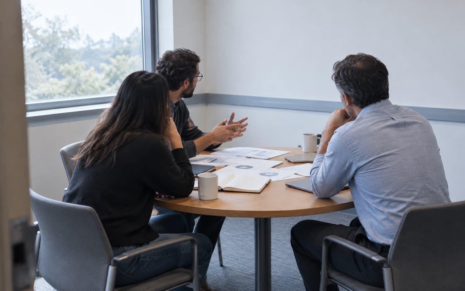 Three people in conversation around a small round table in a plain meeting room, seen from behind, with printed charts and notes spread between them.